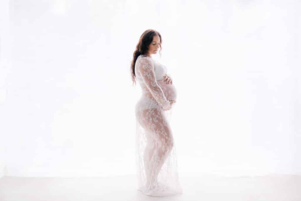 Pregnant woman in white lace maternity gown posing against bright white background in elegant studio photo.