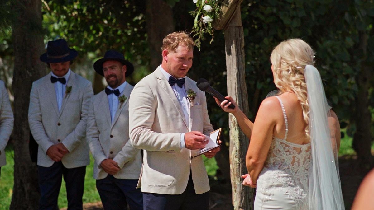 Groom reads vows to blonde bride at outdoor wedding ceremony, groomsmen in bow ties stand behind.