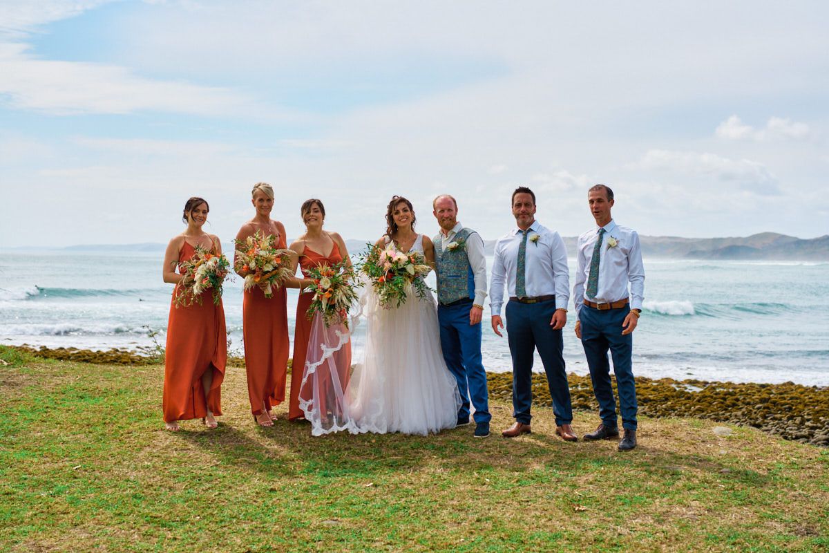 Wedding party posing by ocean, bridesmaids in rust dresses, bride in white gown, groomsmen in blue suits on grassy coastal lawn.