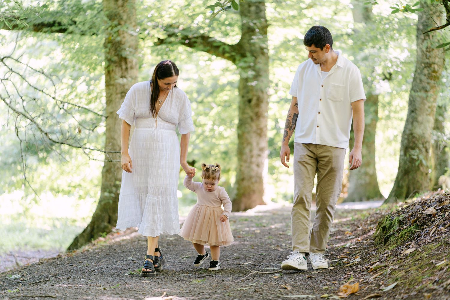 A family of three walks on a forest path, with parents holding their toddler daughter's hands.