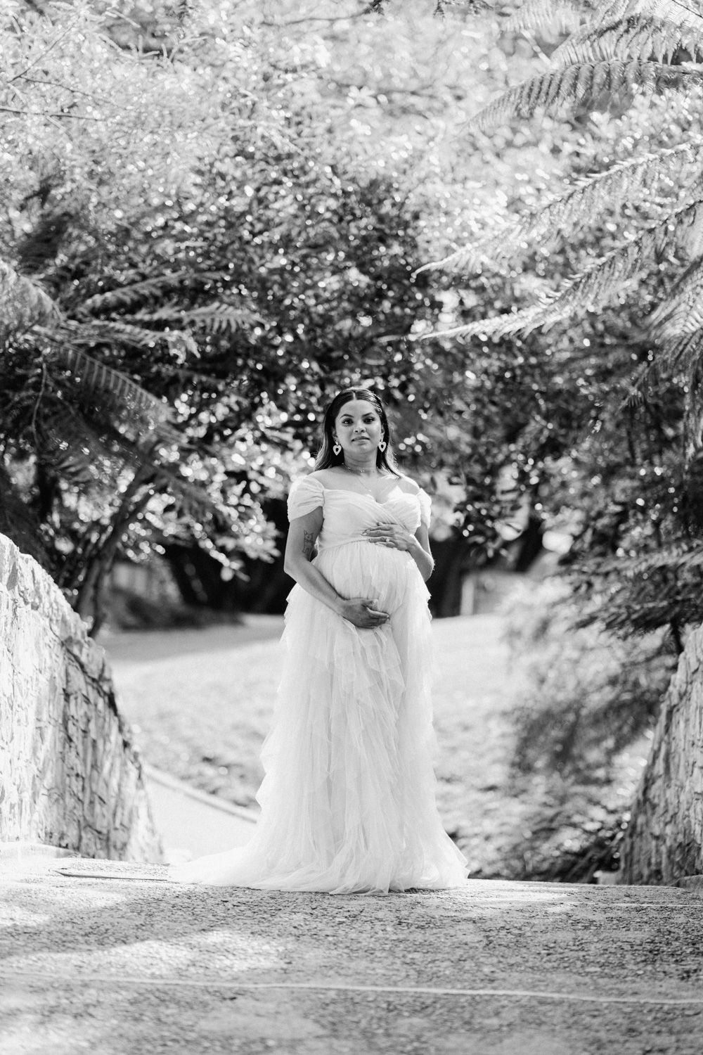 Black and white photo of a pregnant woman in a white tulle gown cradling her belly outdoors among lush trees.