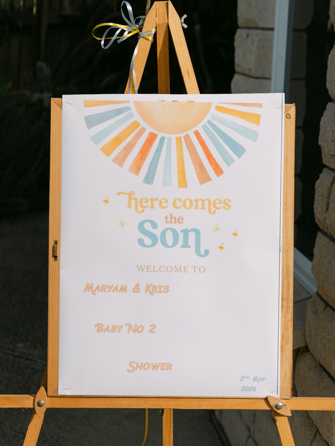 Baby shower welcome sign on easel reading Here Comes the Son for Maryam and Kris Baby No 2 with sun design.