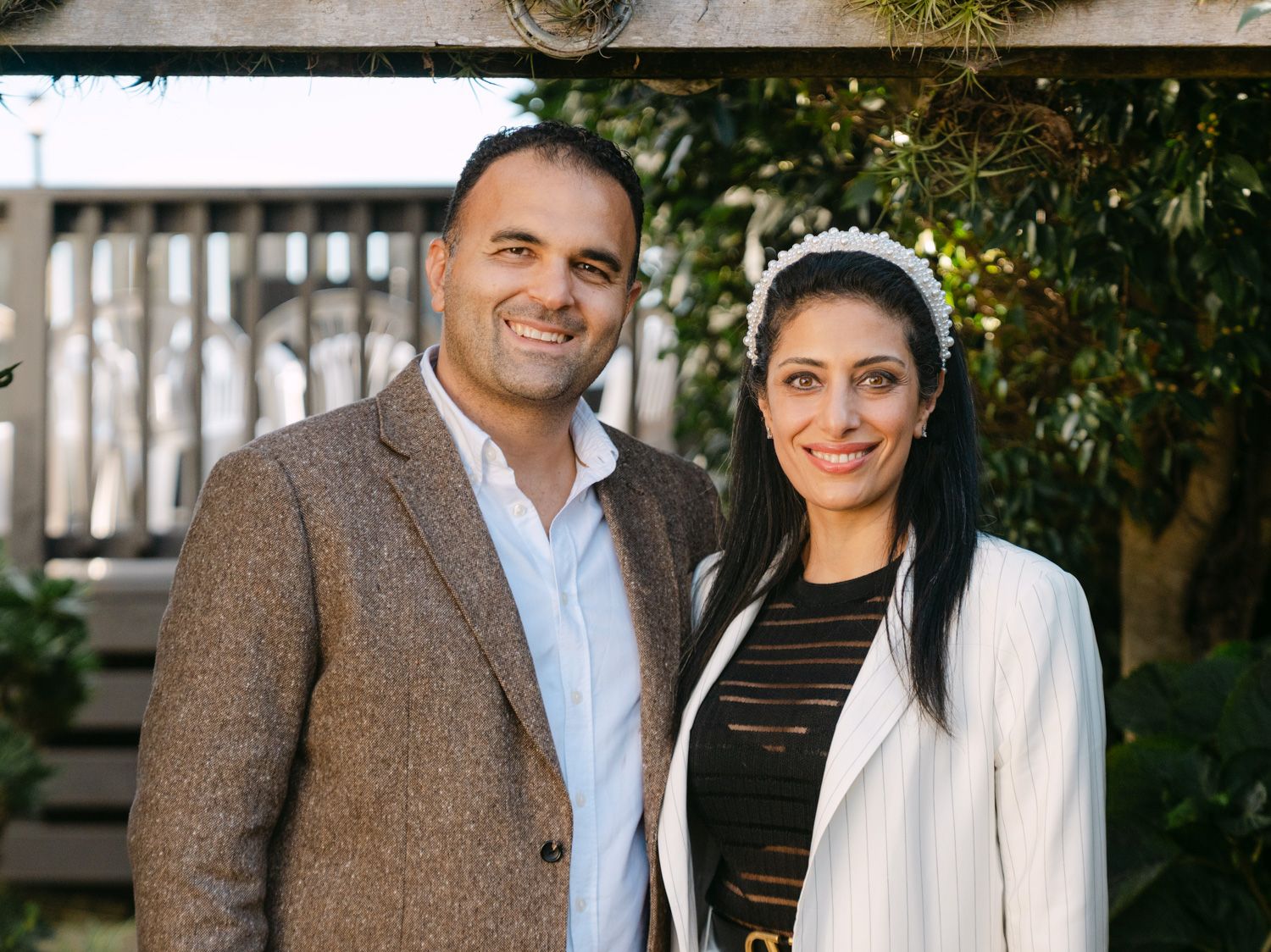 A smiling couple posing outdoors, man in brown blazer and woman in white jacket with floral headband.
