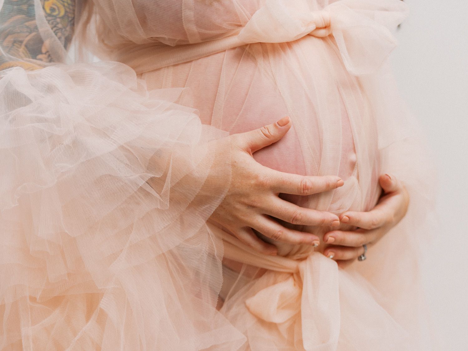 Pregnant woman in a soft pink tulle dress gently cradling her baby bump with both hands.