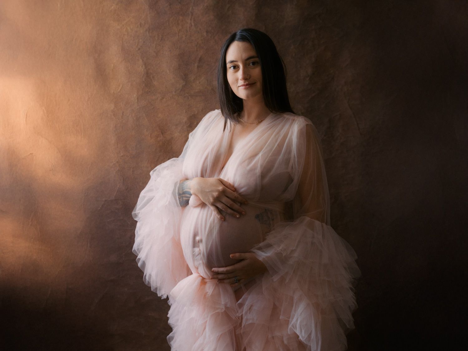 Pregnant woman in pink tulle robe cradling baby bump against brown studio backdrop.