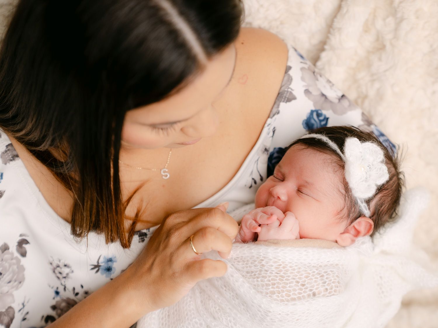 A mother gazes lovingly at her newborn baby wrapped in white, wearing a floral headband, in a soft natural light setting.
