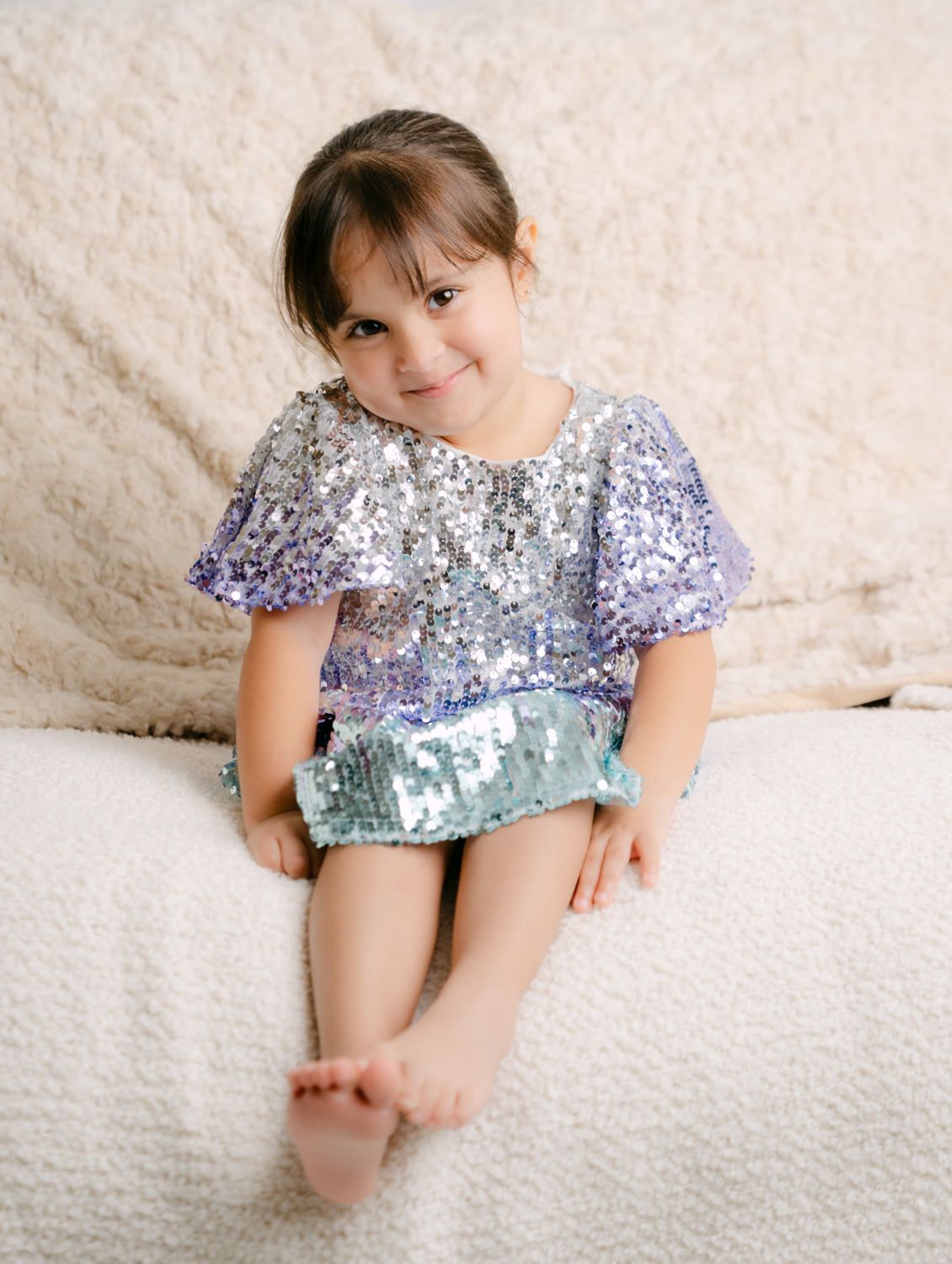 Young girl in a sparkling sequined purple and mint dress sitting barefoot on a cream textured surface, smiling softly.