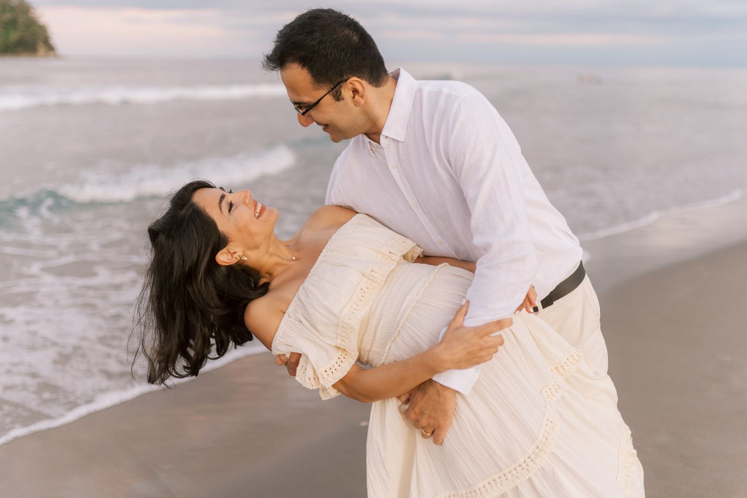 Couple sharing a romantic dip on a sandy beach, woman in white dress laughing as man in white shirt holds her.