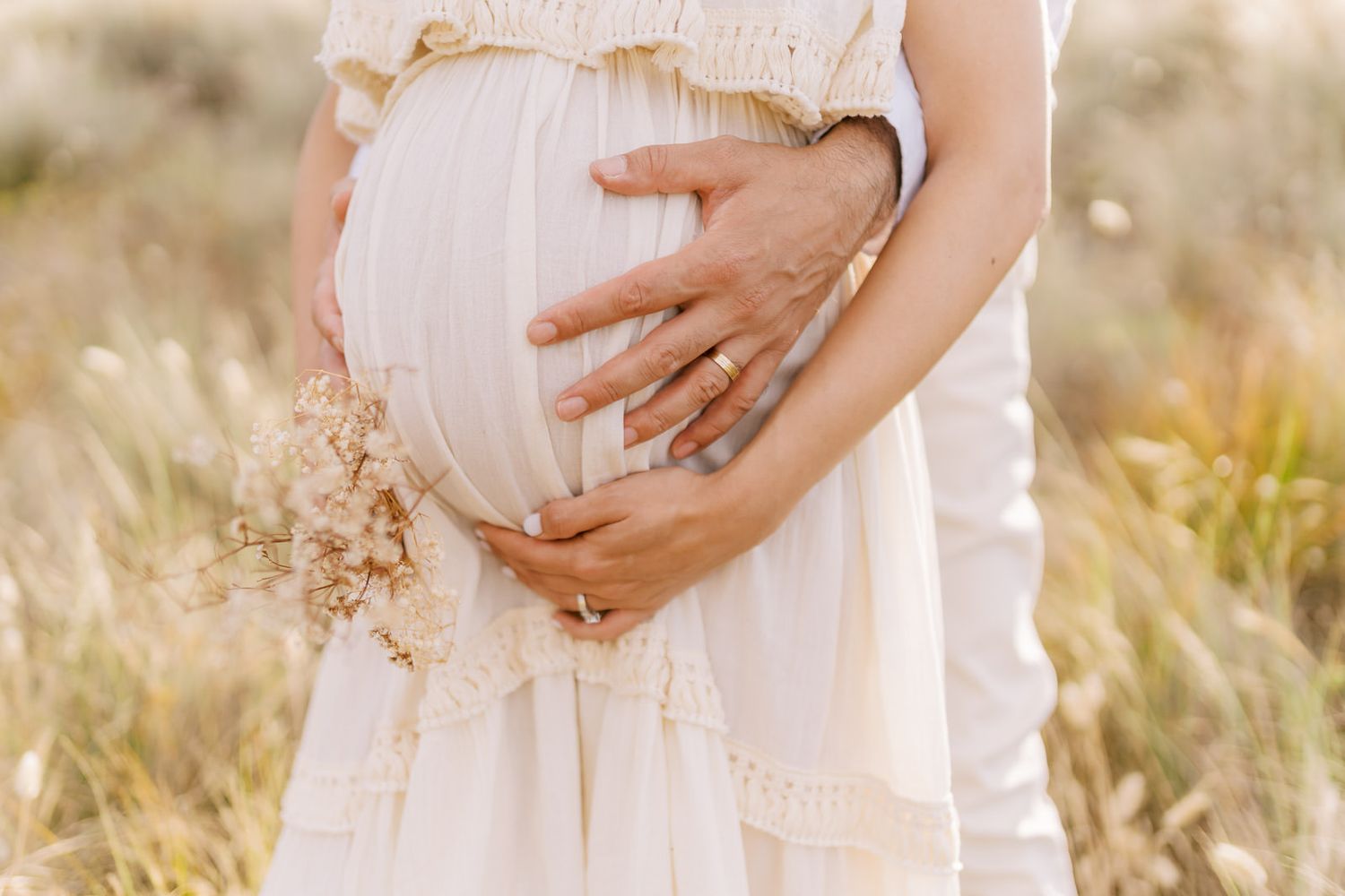 Pregnant woman in white dress with partner's hands cradling baby bump in a golden field.