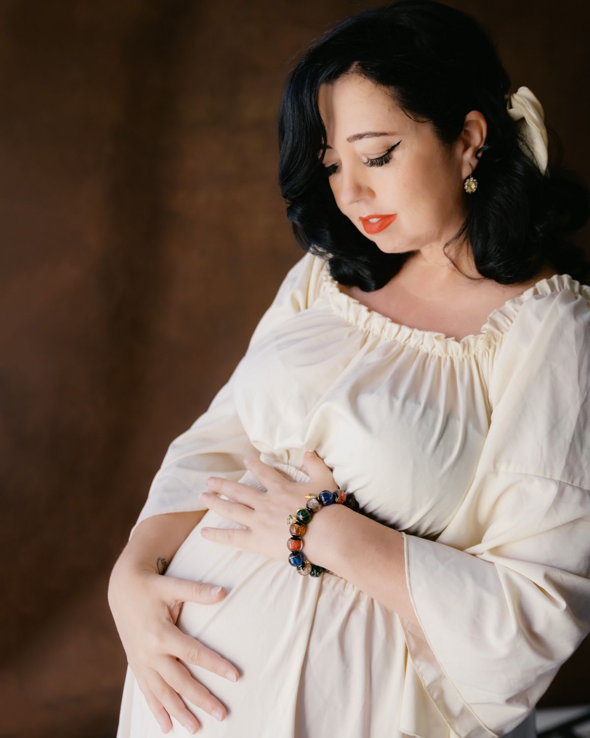 Pregnant woman in cream dress cradling baby bump, wearing colorful beaded bracelet, studio maternity portrait.