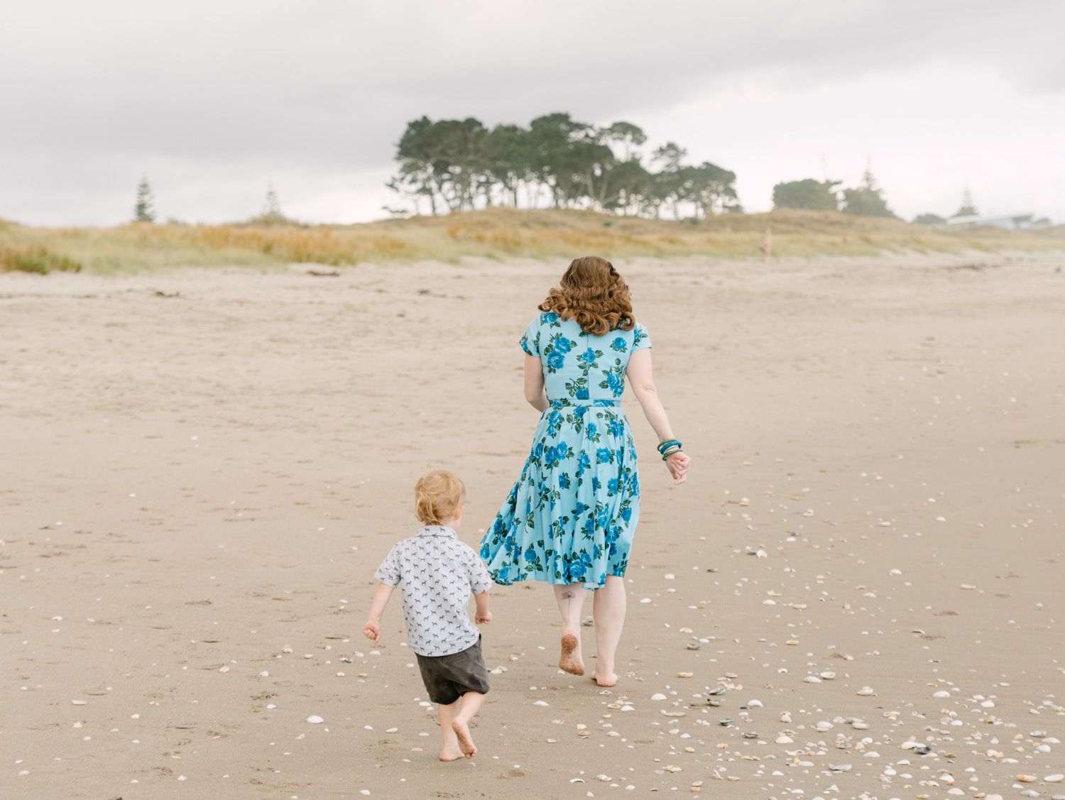 A mother and young child walk hand-in-hand barefoot on a sandy beach, with trees and overcast sky in the background.