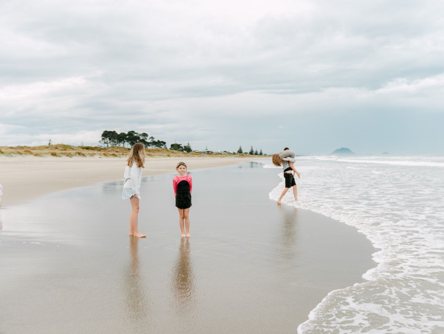Three children playing on a sandy beach near ocean waves under a cloudy sky.