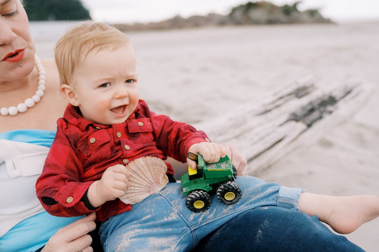 A happy baby boy in a red shirt plays with a John Deere toy tractor on a beach while sitting on an adults lap.