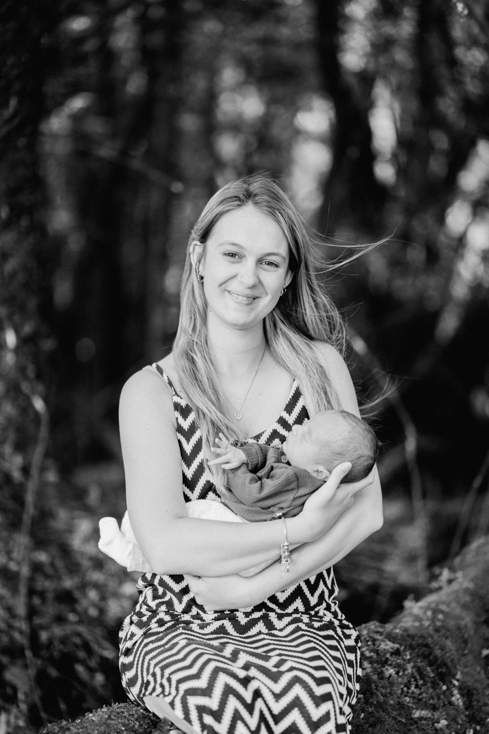 Black and white portrait of a smiling young mother holding her newborn baby outdoors in a forest setting.