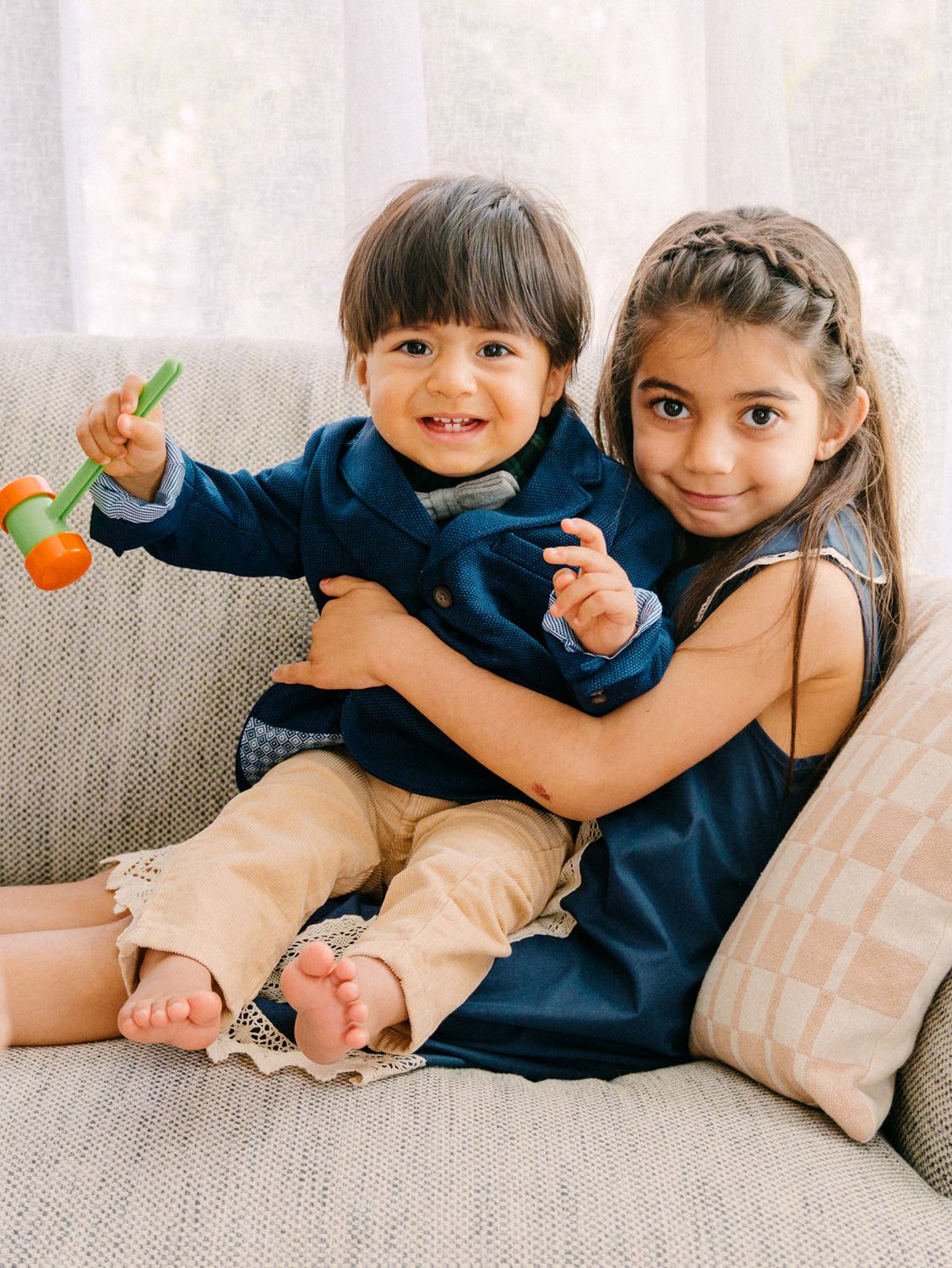 Two young siblings sitting on a cozy sofa, older girl hugging toddler boy holding a colorful toy, both dressed in navy outfits.