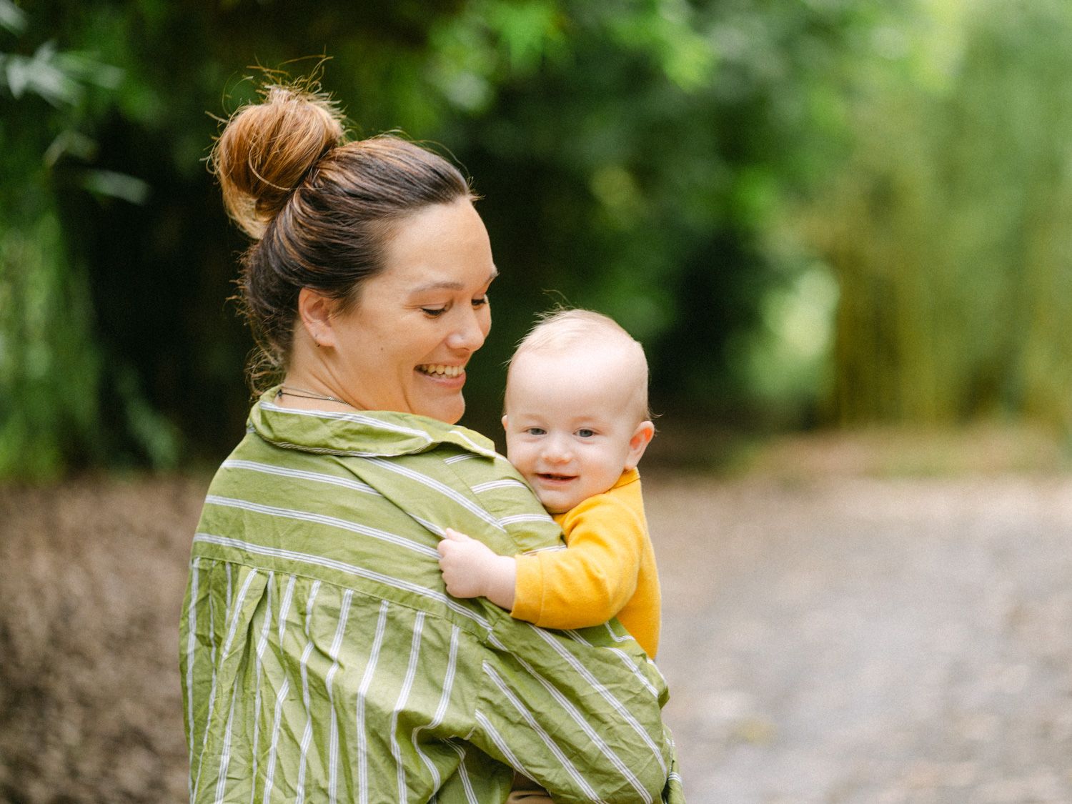Mother carrying smiling baby in green striped wrap carrier outdoors on a nature path.