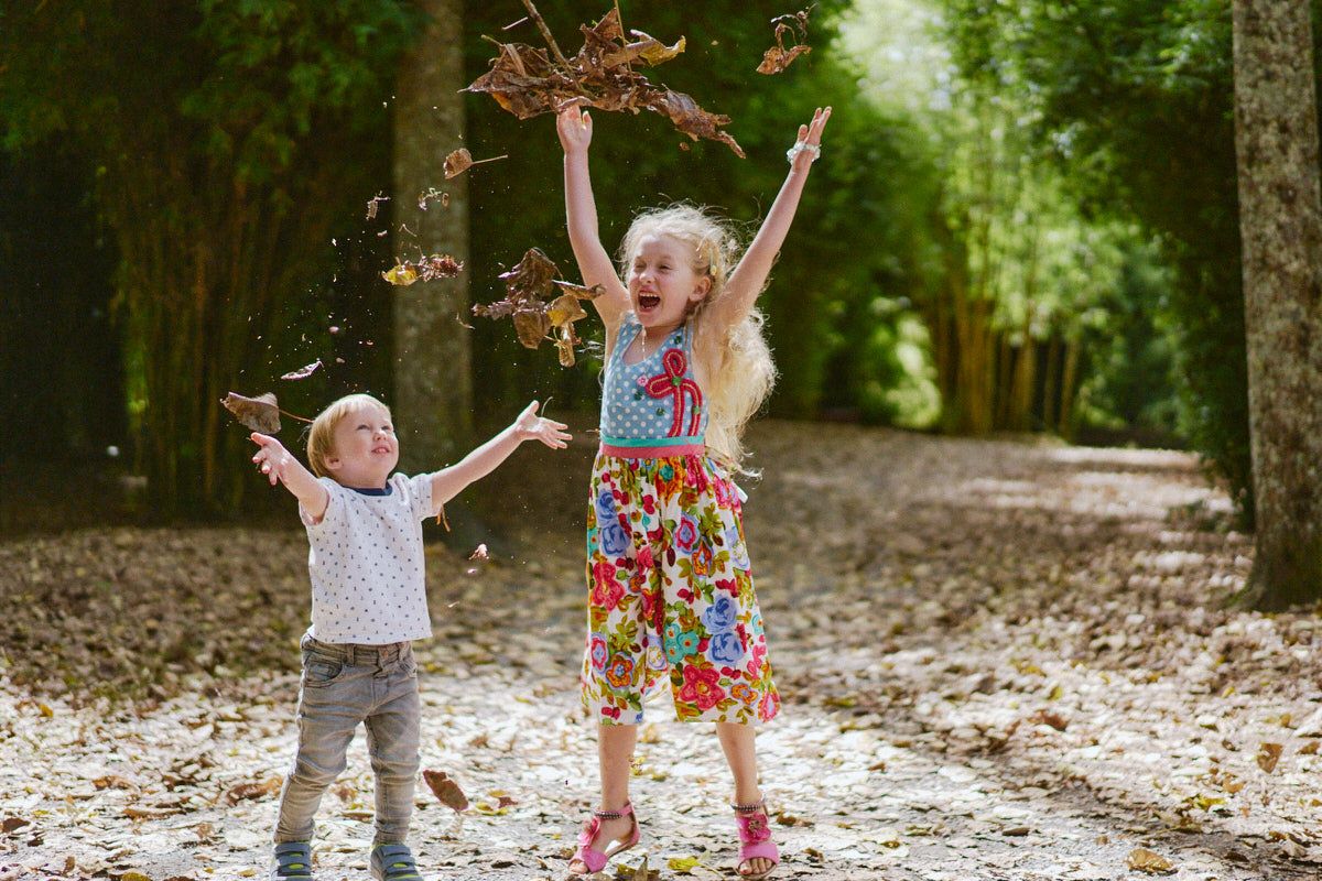 Two joyful children tossing autumn leaves in a tree-lined path, girl in floral dress and boy in casual outfit playing outdoors.