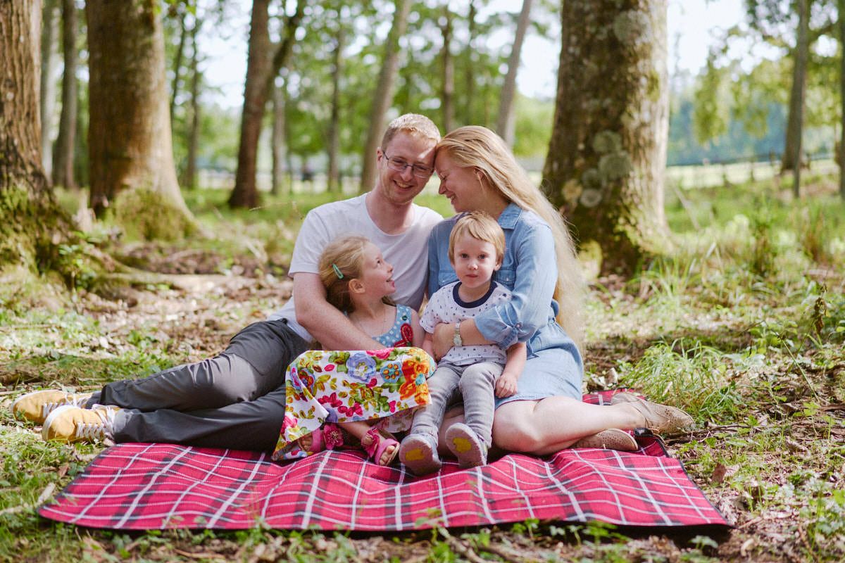 Family of four enjoying a picnic on a red blanket in a wooded park, with two young children sitting together.