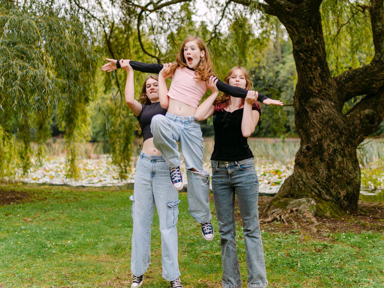 Three girls laughing outdoors, two lifting a third in the air near a willow tree by a pond.