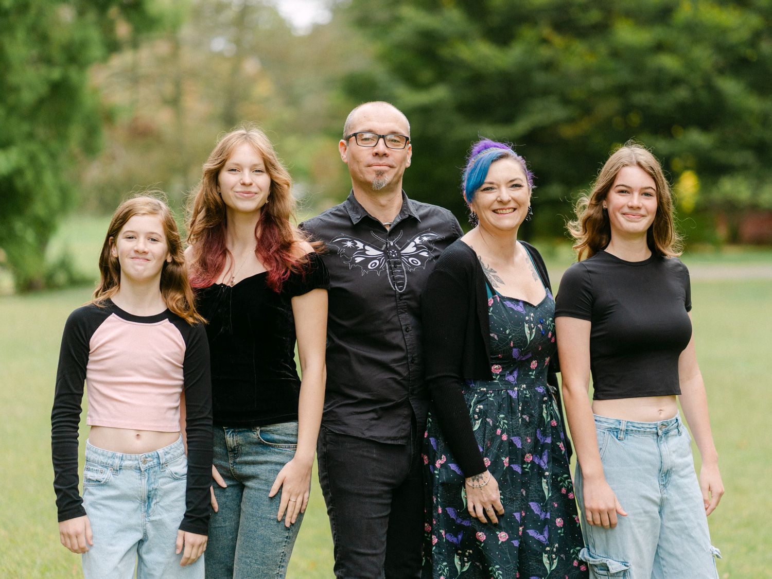 Family of five posing outdoors in a lush green park, smiling together for a portrait photo.