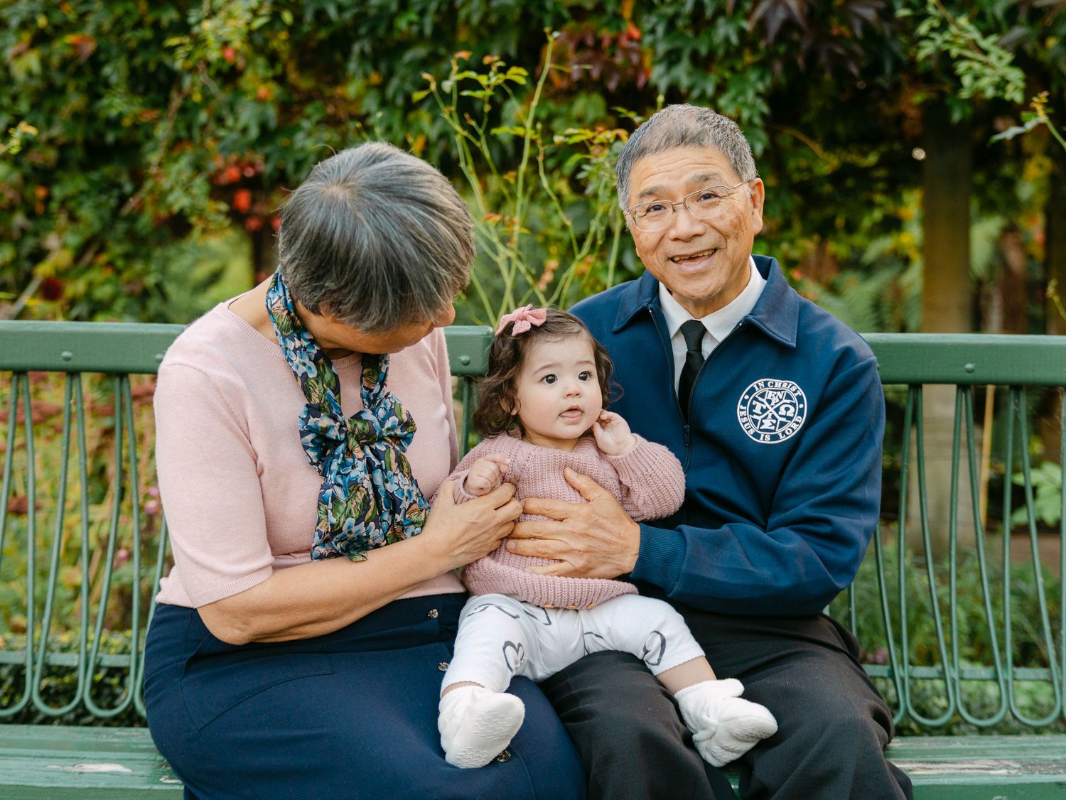 Grandparents sitting on a bench outdoors, lovingly holding a baby girl surrounded by lush green foliage.