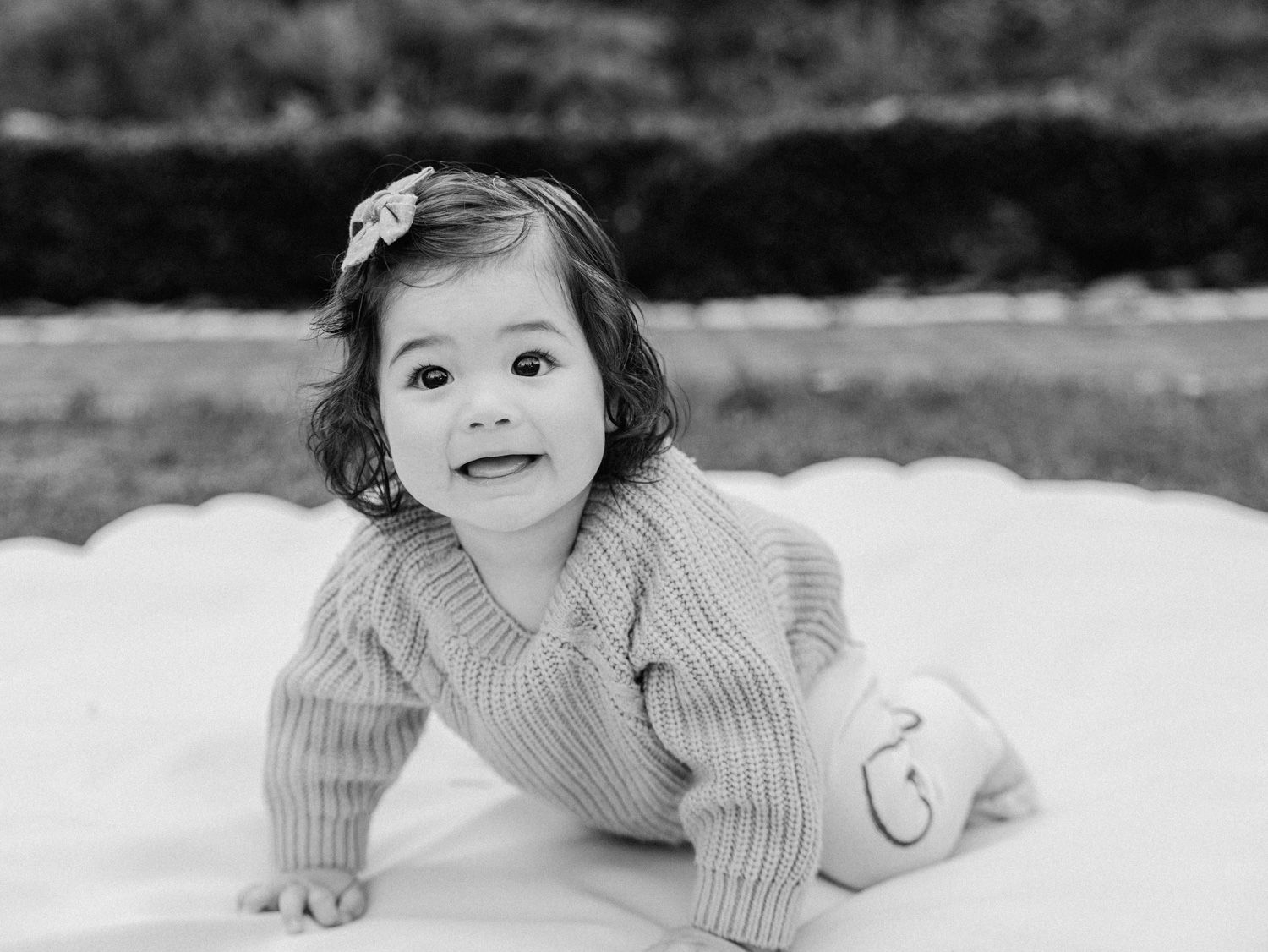 Black and white photo of a smiling baby girl in a knit sweater crawling on a white blanket outdoors.