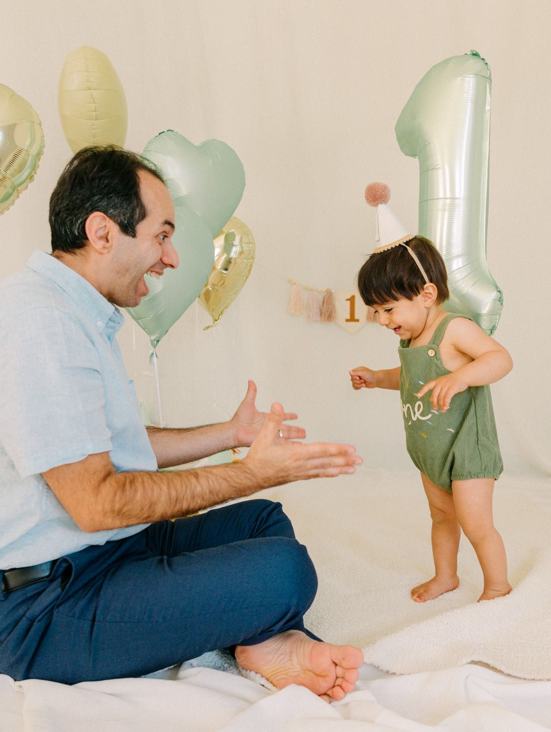 Father claps encouragingly as toddler in green romper and party hat stands at first birthday celebration.