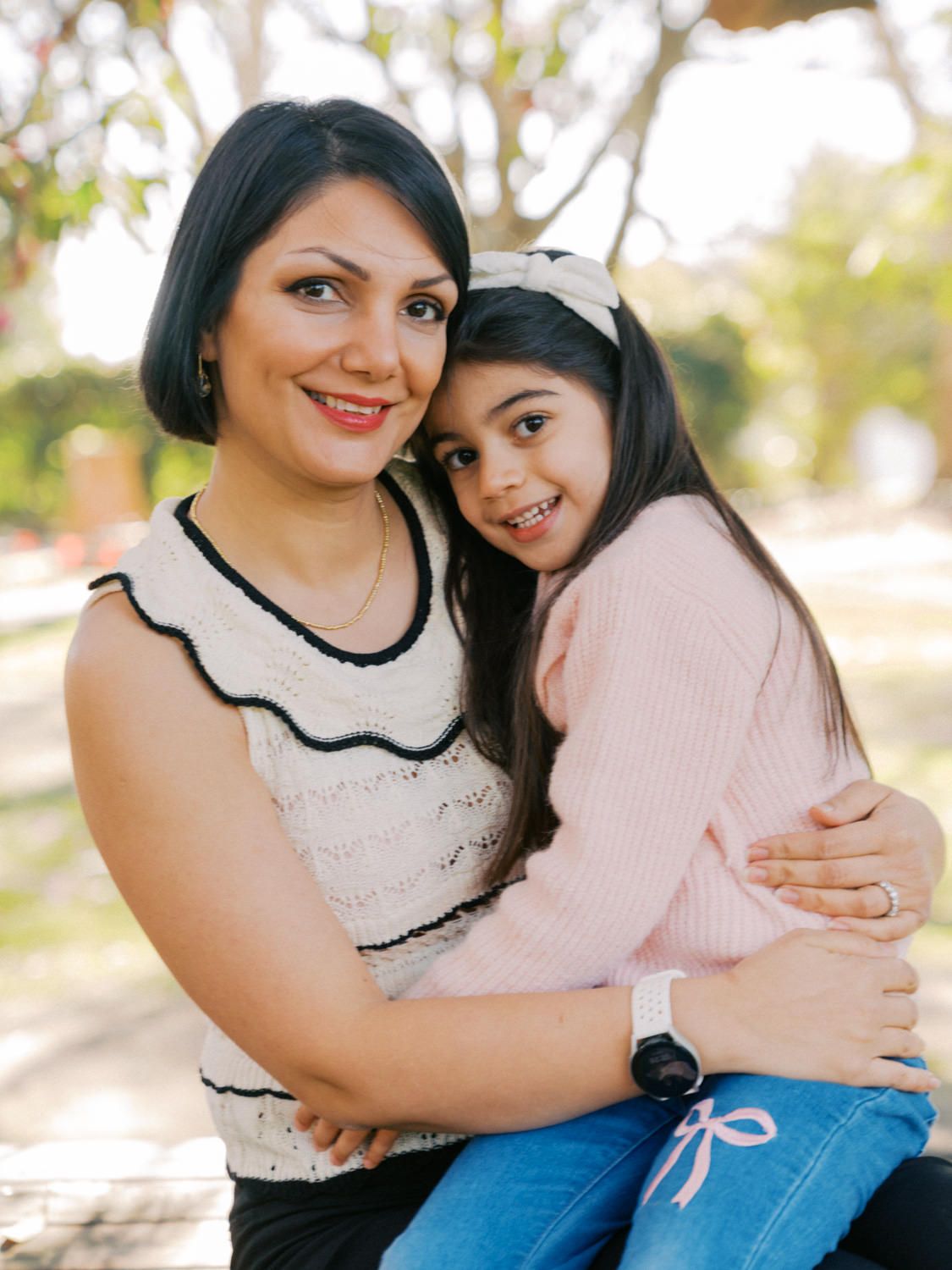 A woman and young girl share a warm embrace outdoors, smiling together in a park setting.