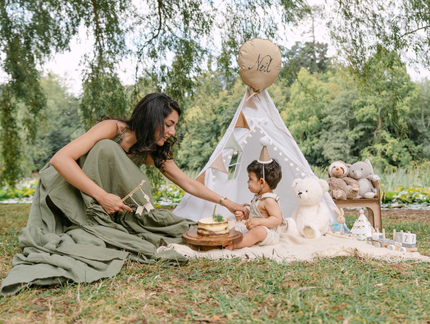 Mother and toddler celebrate first birthday outdoors with a boho teepee, teddy bear, and picnic setup in a lush green park.
