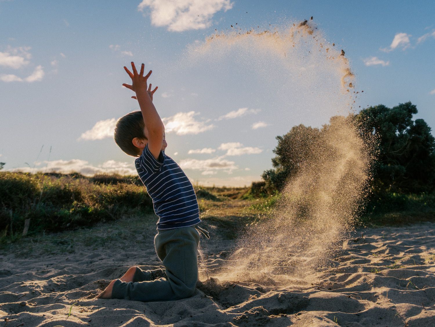 A young boy kneeling on sandy beach tossing sand into the air against a blue sky.