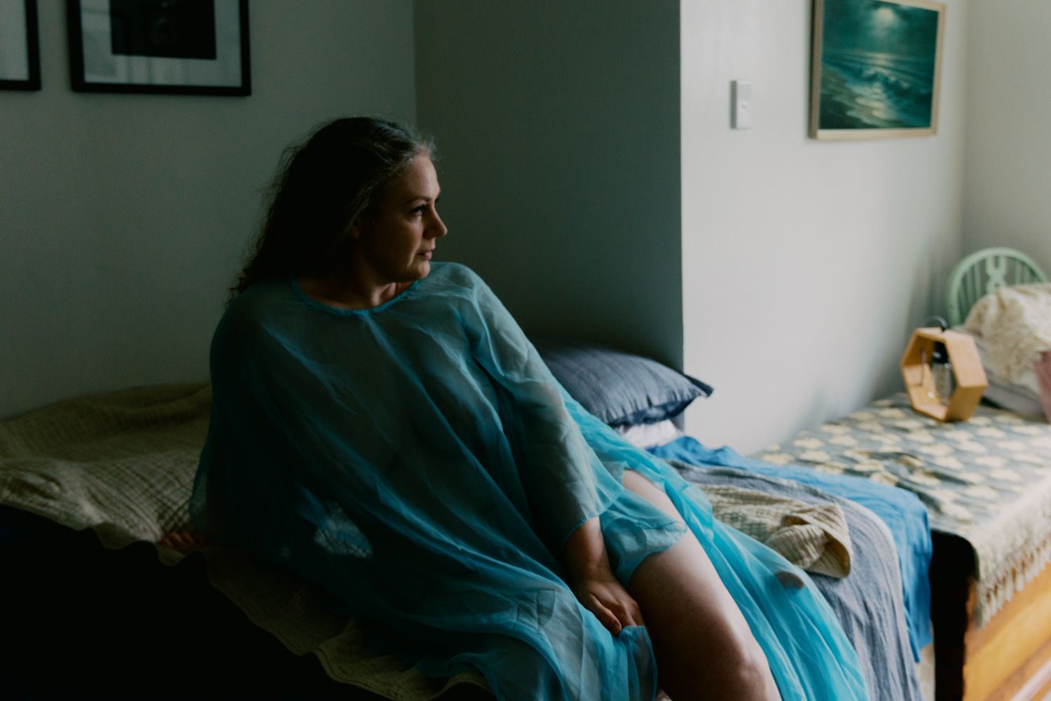 Woman in blue robe sitting pensively on bed near window in softly lit bedroom.