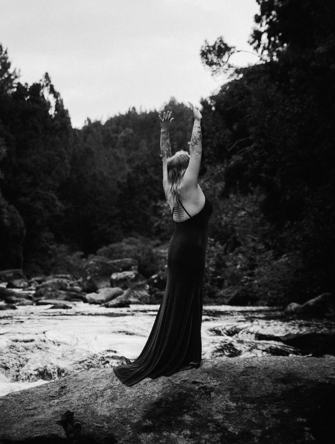Woman in black gown raises arms skyward on rocky riverbank surrounded by lush forest in black and white.