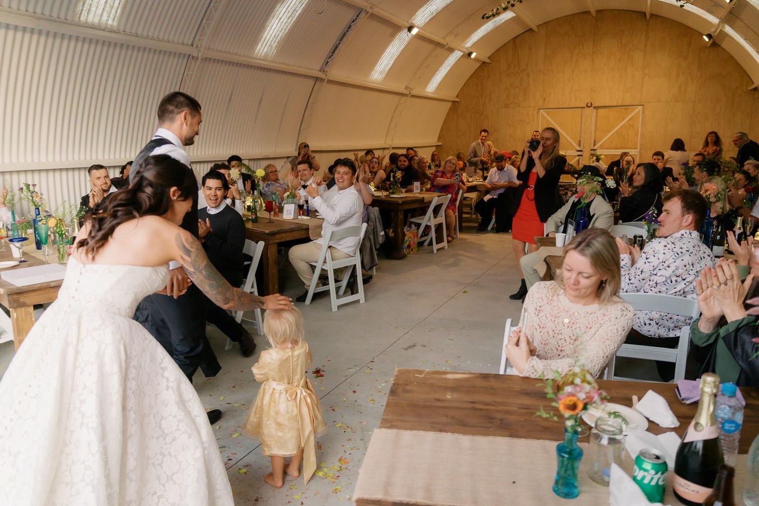 Bride and groom embracing guests at a rustic wedding reception inside a barn venue with seated attendees.