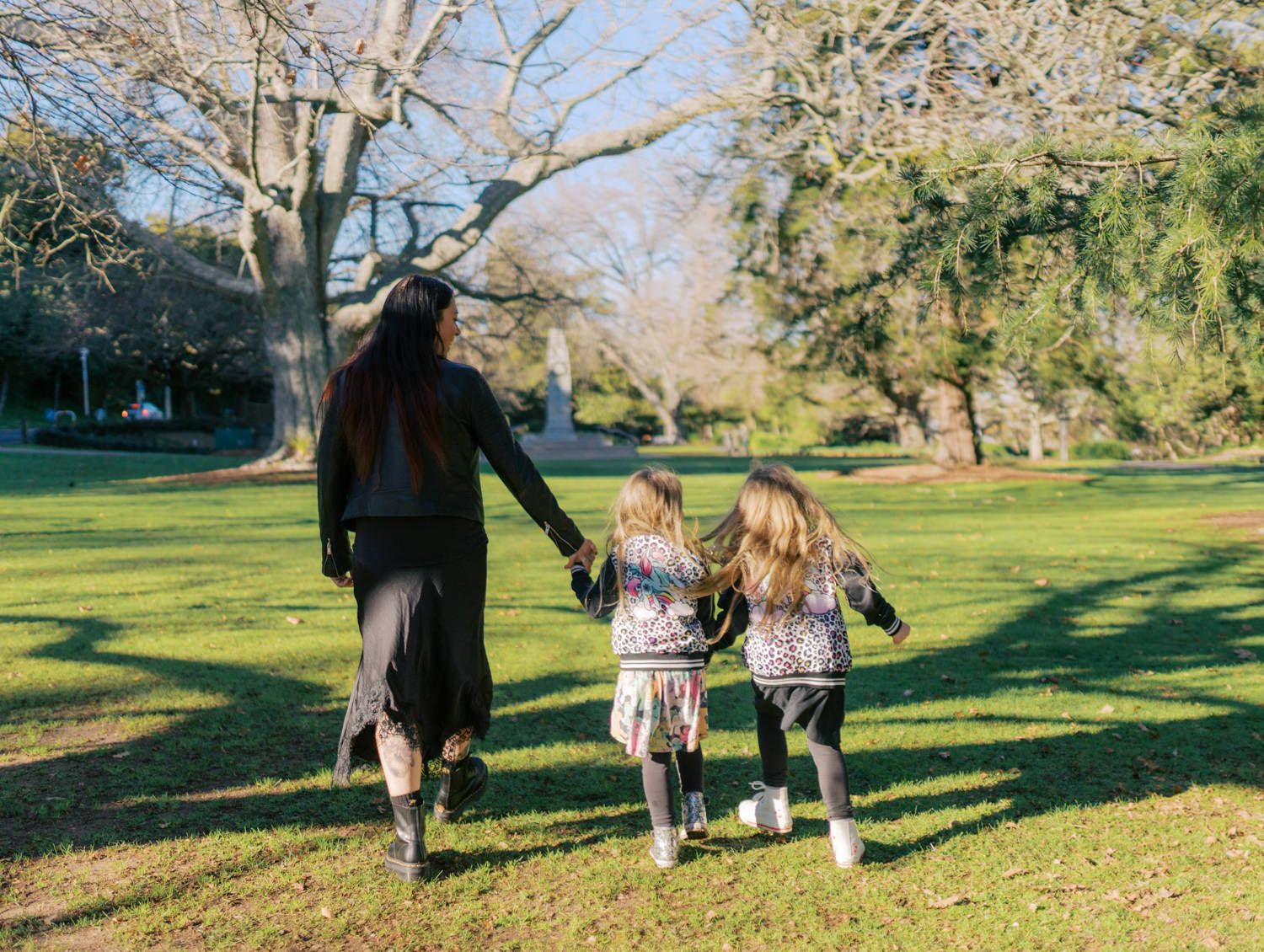 A mother walks hand-in-hand with two young girls across a lush green park on a sunny day.