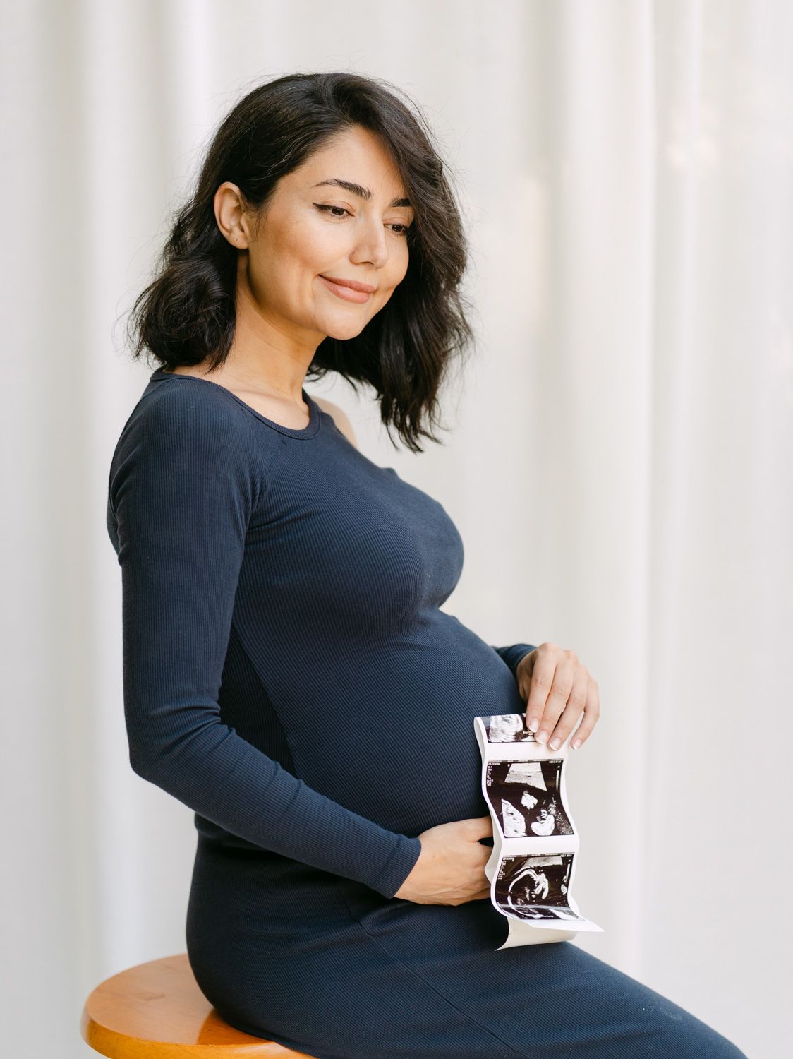 Pregnant woman in navy dress holding ultrasound photos while sitting, cradling baby bump with gentle smile.