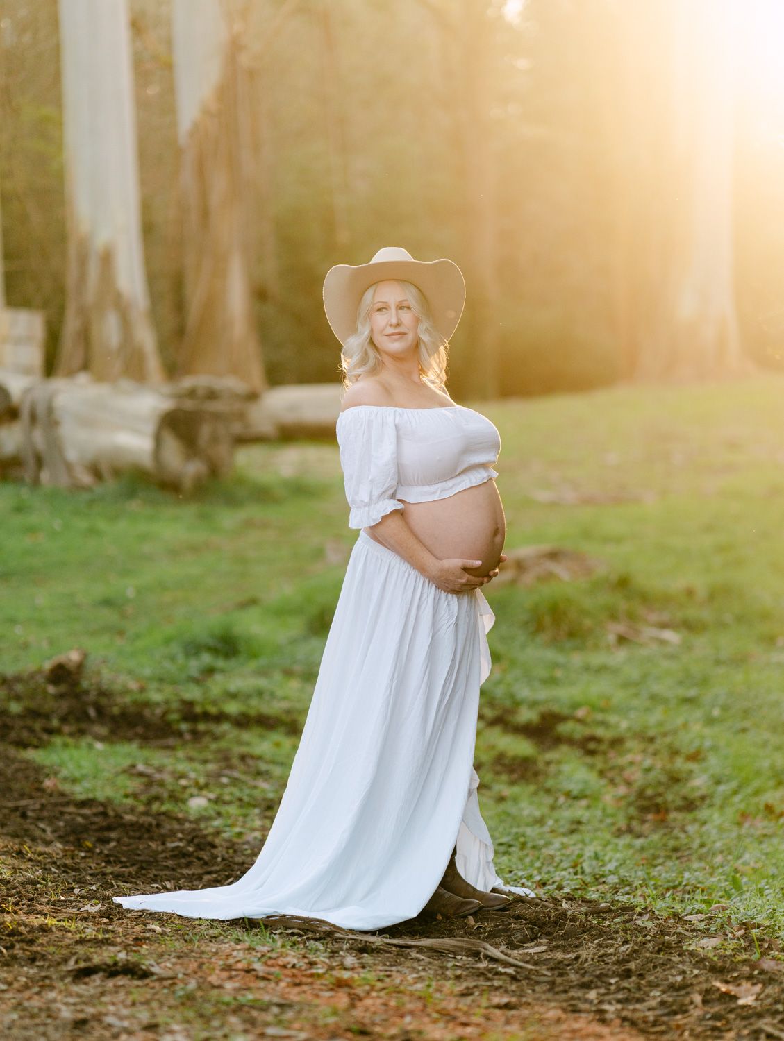 Pregnant woman in white off-shoulder dress and wide-brim hat cradling baby bump in sunlit forest clearing.