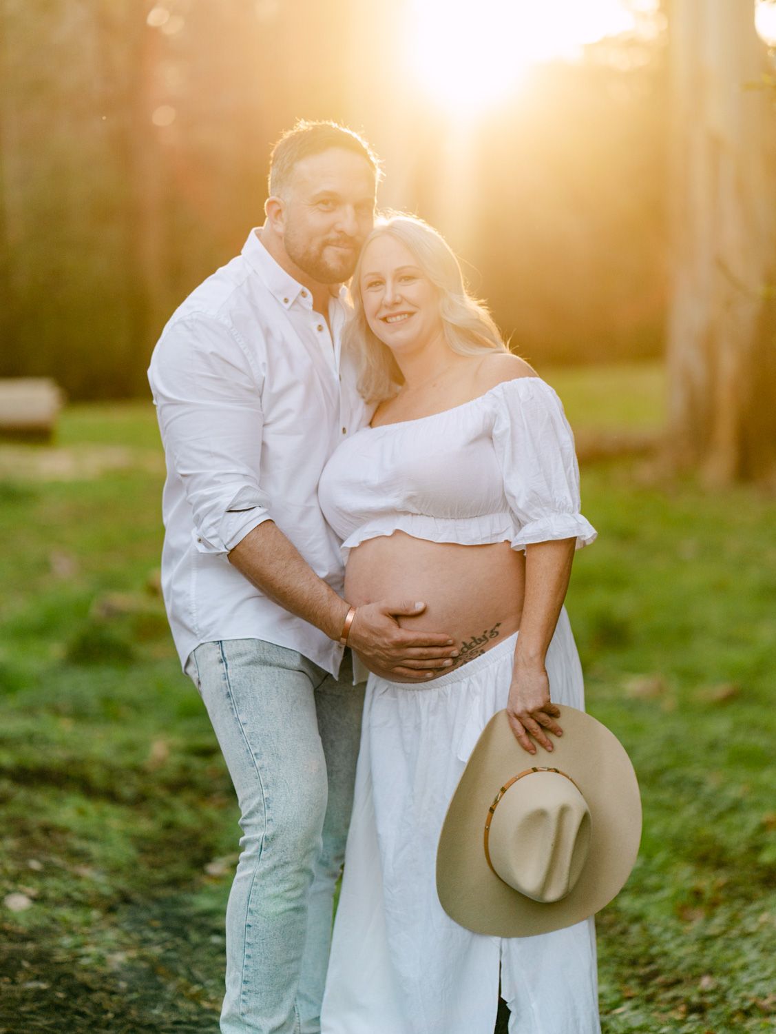 Couple in white outfits poses for golden hour maternity photo in a sunlit park, woman holding a tan hat.