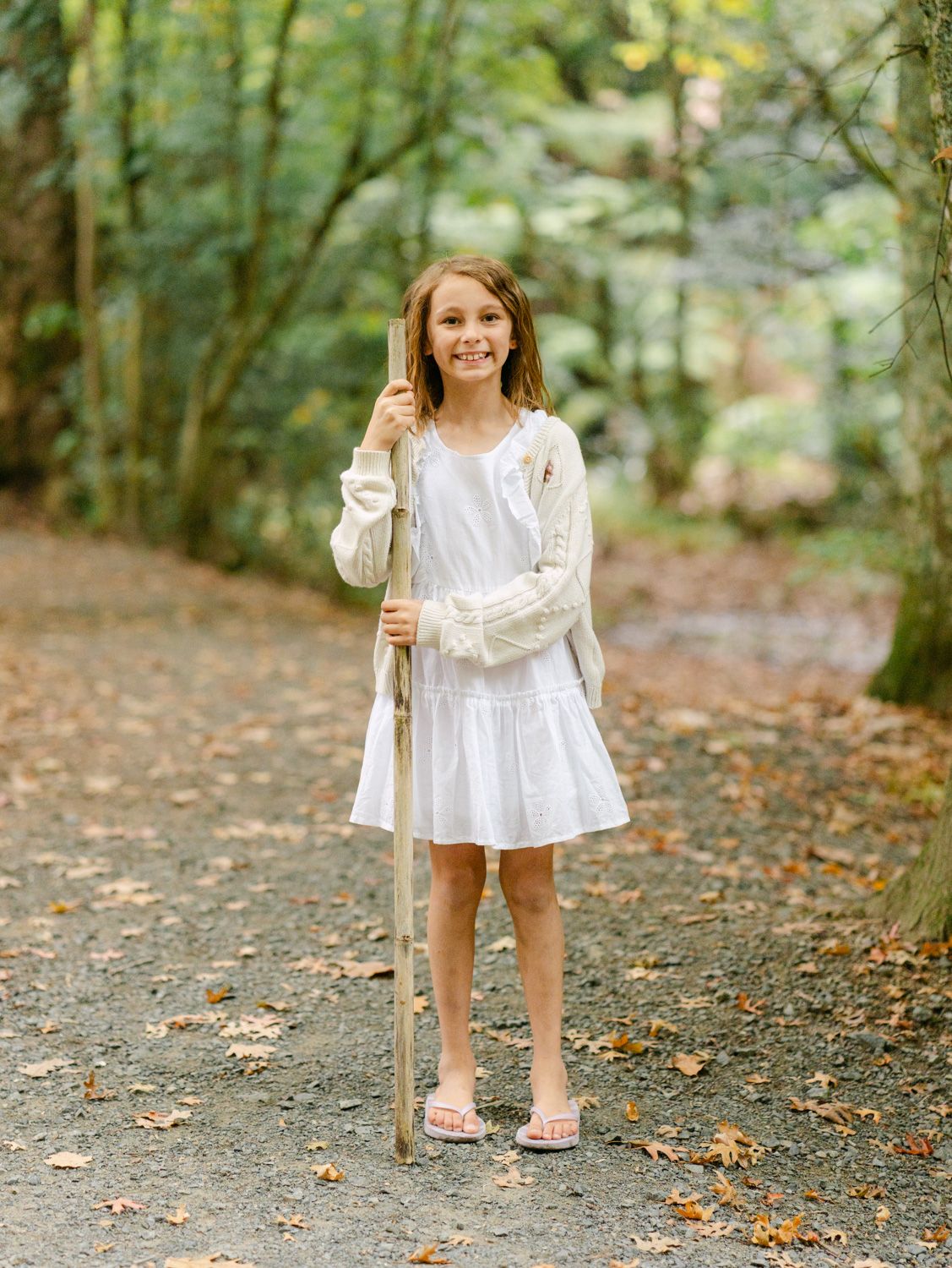 A smiling girl in a white dress and cardigan holds a wooden walking stick on a forest path surrounded by autumn foliage.