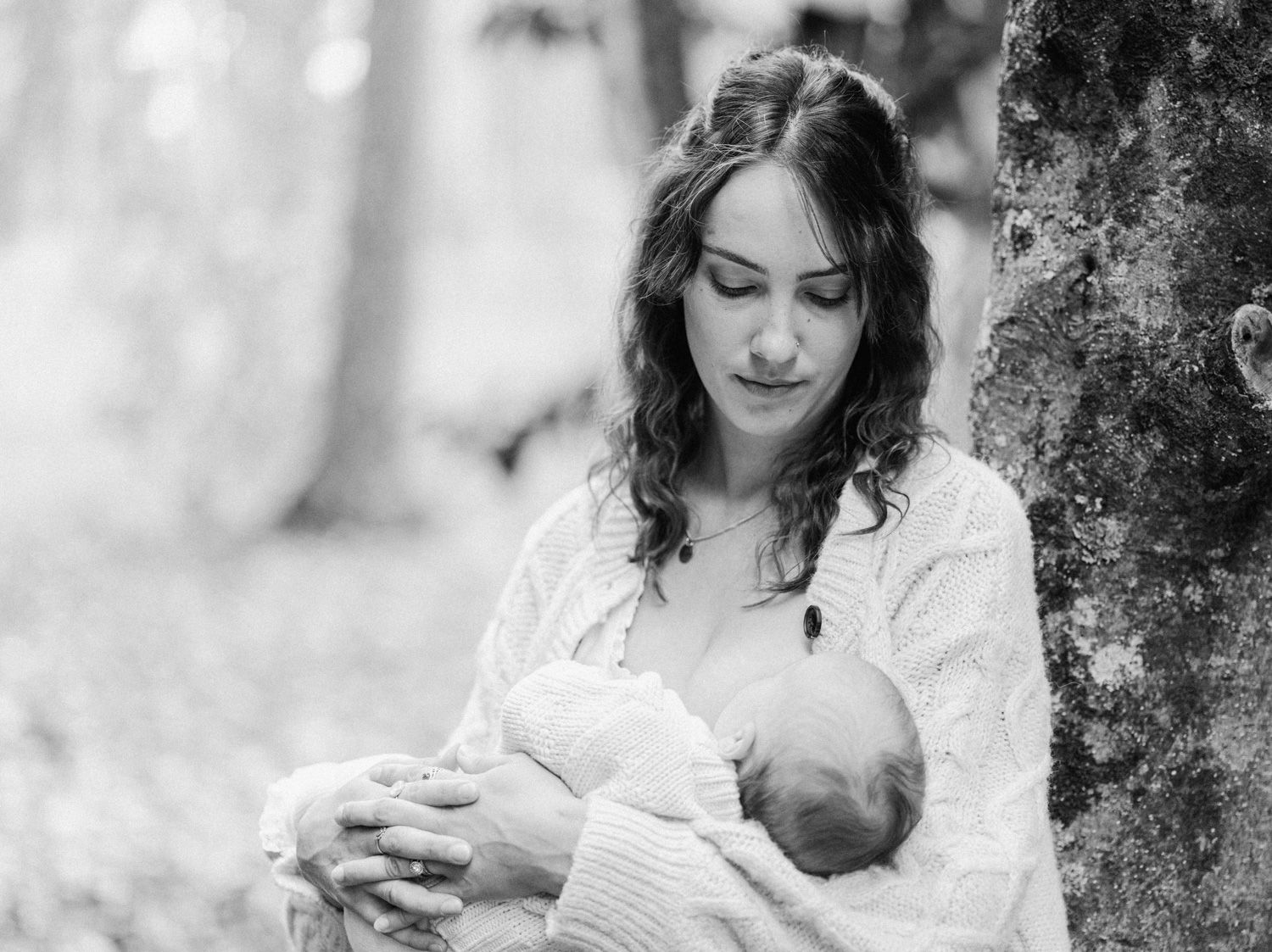 Black and white photo of a young mother tenderly holding her newborn baby outdoors near a tree.