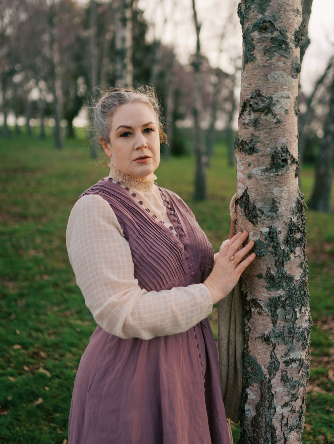 Woman in purple vintage dress leaning against birch tree in autumn park setting.