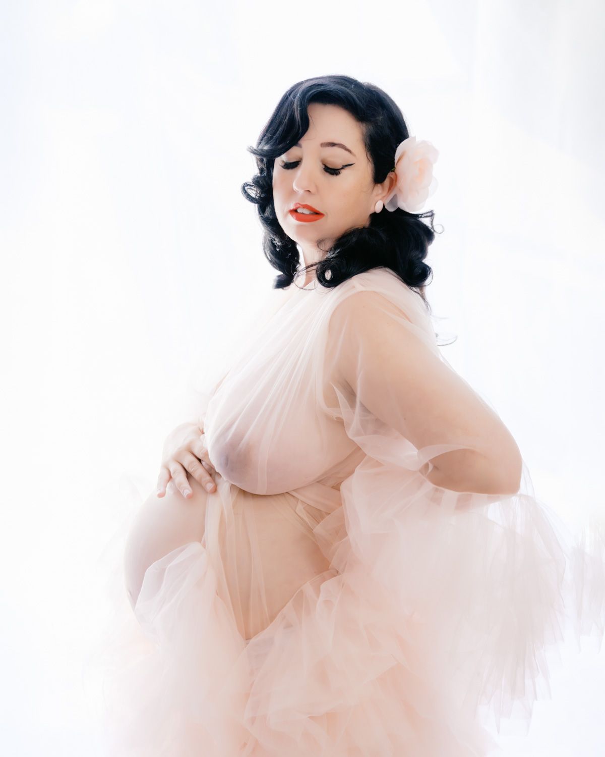 Pregnant woman in sheer blush pink tulle gown with floral hair accessory poses elegantly against white background.