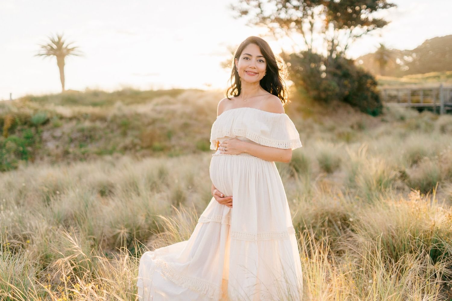 Pregnant woman in white off-shoulder gown standing in golden field at sunset, cradling baby bump.