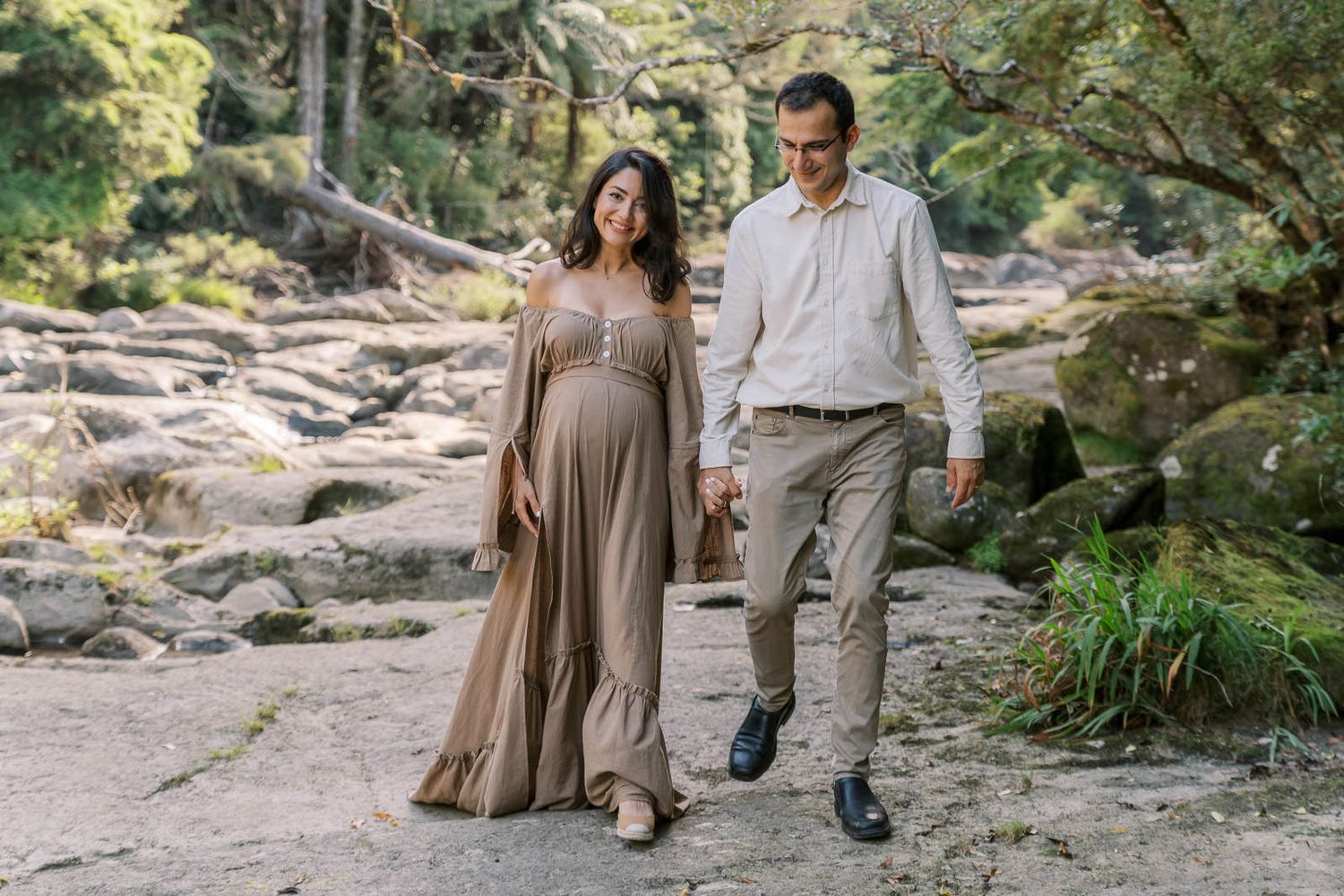 Pregnant woman in flowing tan dress walks hand-in-hand with man on rocky forest path during maternity photoshoot.