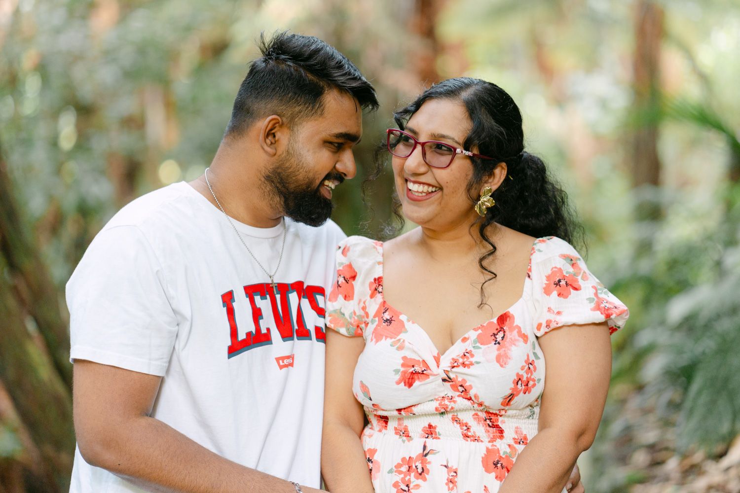 Smiling couple in a lush green forest, man in Levis shirt, woman in floral dress, sharing a joyful moment.