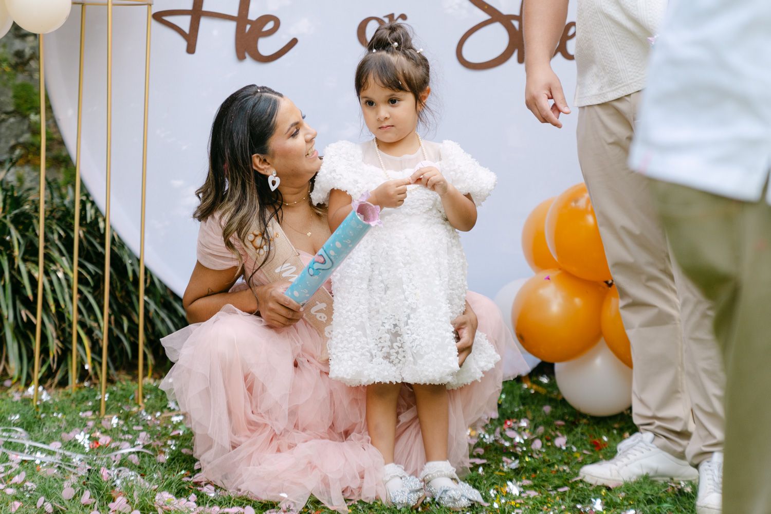 Woman in pink dress crouches beside toddler girl at a gender reveal party with balloons and confetti outdoors.