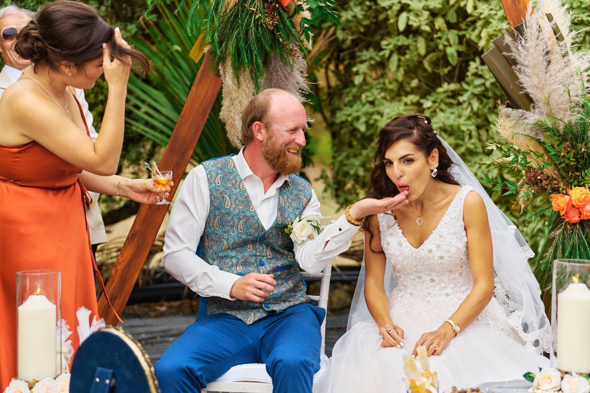 Bride blowing a kiss while groom smiles at outdoor wedding reception with tropical decor and candles.