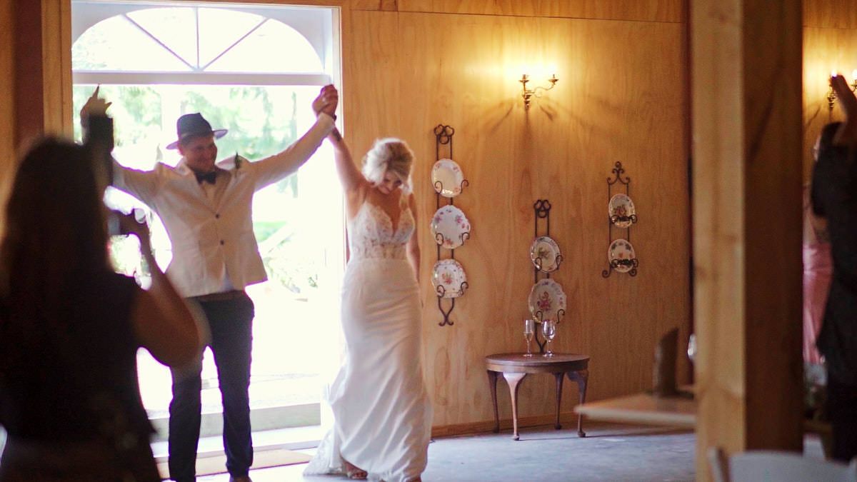 Newlywed couple entering wedding reception hall holding hands raised, bride in white gown, guests celebrating indoors.