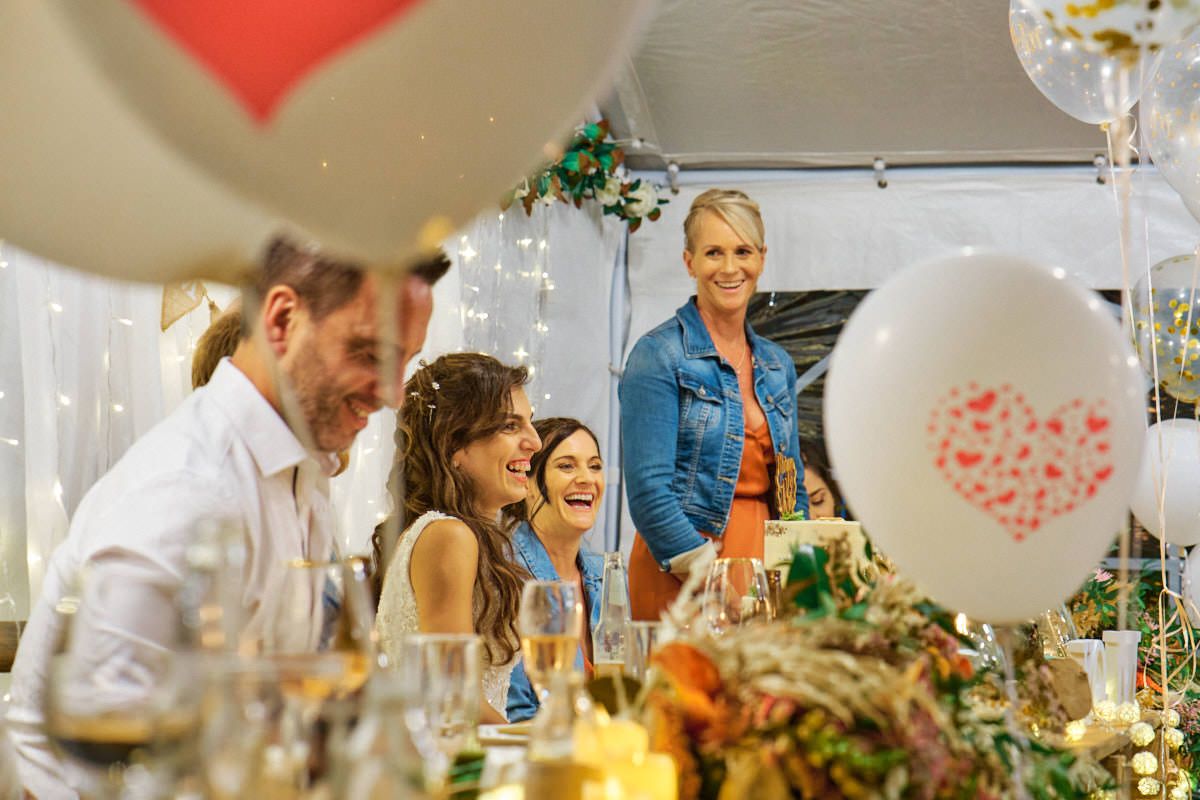 Group of friends laughing at festive party table with heart balloons and fairy lights in decorated tent.
