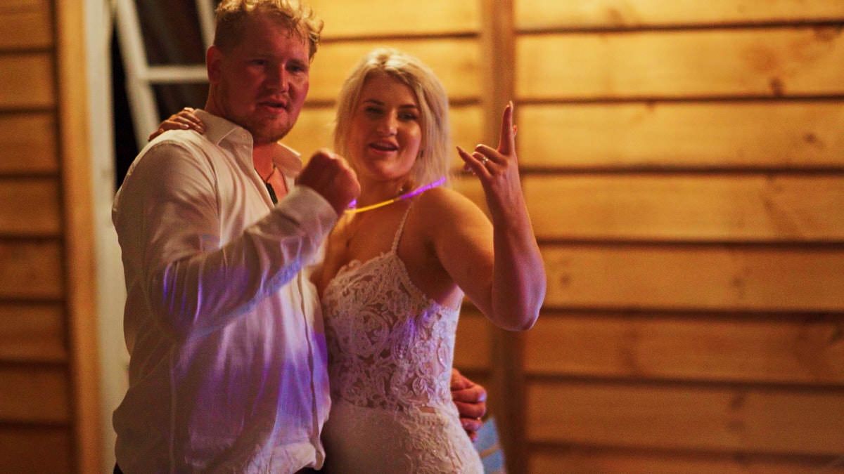 Couple dancing together at an indoor party, woman in white lace dress, man in white shirt, wooden wall background.