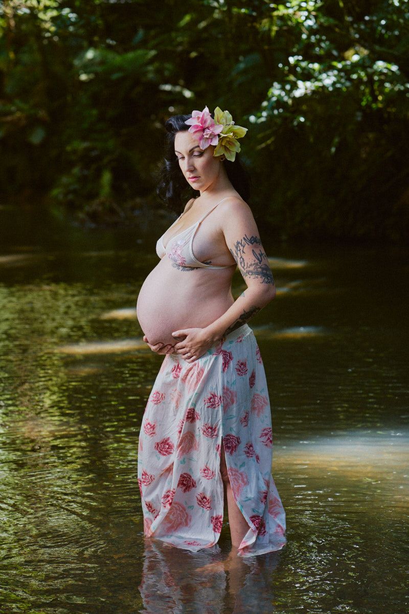 Pregnant woman in floral skirt and flower crown stands in a serene forest stream, cradling her baby bump.